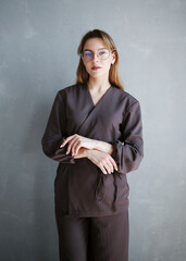 Portrait of a young woman in brown medical scrubs and eyeglasses standing against a gray wall, looking confident and professional.