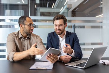 Two businessmen in a modern office setting collaborate, reviewing data on a tablet and laptop,...