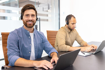 Two customer service representatives wearing headsets sit at a table with laptops, smiling and...