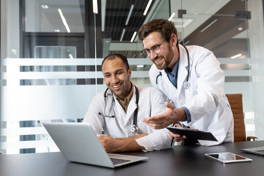 Two doctors, smiling, look at a laptop screen during a discussion in a modern office setting.