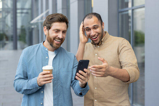Two diverse men are looking at a phone, smiling and happy. One is holding coffee.