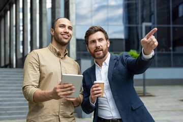 Two businessmen in front of a modern building discussing a project, one pointing the other holding a tablet.