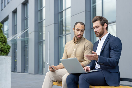 Two business professionals collaborating outdoors near a modern building, using a laptop and a tablet.