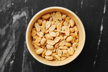 Salted peanuts in a bowl. Delicious snack for party or bar. Tasty roasted nuts on black marble table. Crunchy texture and golden color. Top view of popular food for drinks and celebrations.