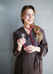 Portrait of a young female doctor in glasses and brown scrubs holding a pink stethoscope and smiling against a light gray wall.