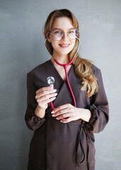 Portrait of a young female doctor in brown scrubs and glasses smiling while holding a pink stethoscope against a light gray wall.