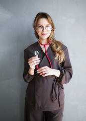 Confident young female doctor with glasses and long hair holding a stethoscope, wearing modern brown scrubs, standing against a light wall.