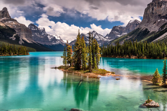 View of a turquoise lake mirroring the majestic, snow-capped mountains and a small island of evergreens under a cloudy sky, Jasper National Park, Alberta, Canada.