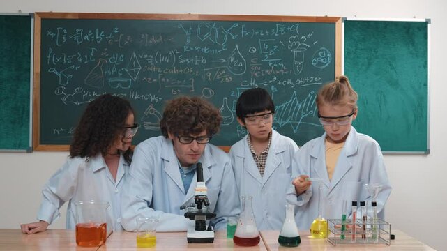 Smart caucasian teacher looking under microscope while diverse children doing experiment at STEM science lesson. Professional happy scientist inspect biochemical example by using microscope. Pedagogy.
