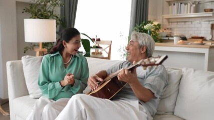 Happy senior man sitting at sofa and playing guitar or music instrument while his wife singing and moving to music. Elder couple spending time together and enjoy doing activity together. Myrmidon. - Powered by Adobe