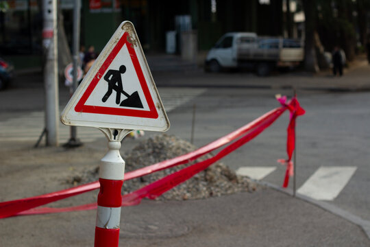 Horizontal street city photo. Road repair sign with black person icon digging ground in white triangle with red outline. Danger unsafe area warning red tape near pedestrian crossing. Cross line. Risk