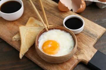 Half-boiled egg in bowl, soy sauce, chopsticks, coffee and toasted bread on wooden table, closeup....