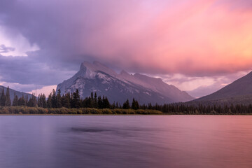View of tranquil waters reflecting the majestic mountain range under a dramatic, cloud-infused sky painted with hues of dawn, Vermilion Lake, Alberta, Canada.