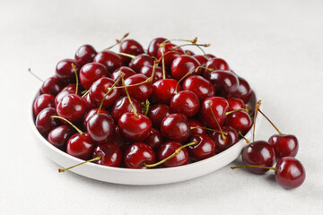 Heap of fresh ripe red sweet cherry with stems in white plate on light textured background. Natural summer fruit, healthy organic food, harvest and seasonal concepts.