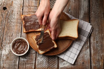 Woman spreading tasty chocolate butter onto bread at wooden table, top view