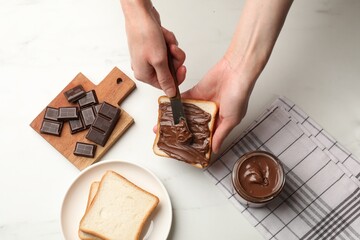Woman spreading tasty chocolate butter onto bread at white table, top view