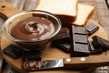 Tasty chocolate butter in glass bowl, pieces, bread and knife on color wooden table, closeup