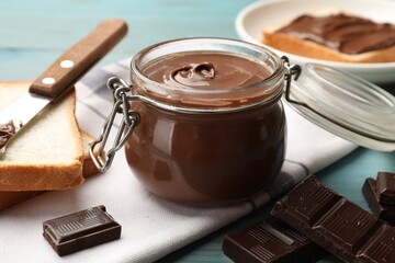 Tasty chocolate butter in jar, pieces, knife and bread on light blue wooden table, closeup