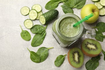 Tasty smoothie in mason jar, spinach leaves, apple, kiwi and cucumber on grey textured table, above...
