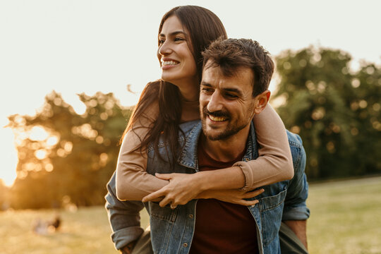 Happy couple enjoying piggyback ride in golden hour sunlight at park - Powered by Adobe