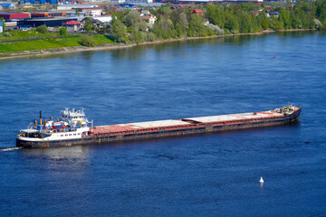 A long cargo ship is seen gliding through the calm waters of a river during spring. Lush greenery...