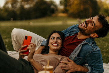Happy couple relaxing using smartphone at park, enjoying picnic