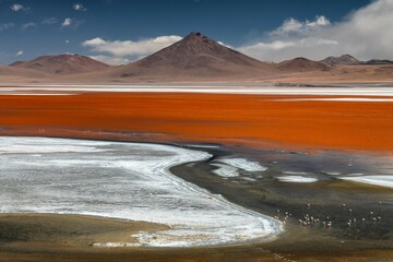 View of vibrant red waters meet the stark white shores under a vast blue sky, a landscape dominated by towering mountains, Potosi Department, Bolivia.