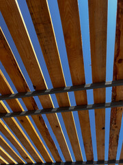Modern wooden pergola roof with evenly spaced slats against a clear blue sky. The architectural structure creates a dynamic geometric pattern with light and shadow, highlighting natural wood texture