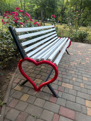 Romantic park bench with heart-shaped armrest surrounded by blooming roses in a peaceful garden. Love and relaxation concept.