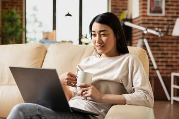 Female freelancer sits with laptop on laps and holds coffee mug, focused on online webinar. Remote...