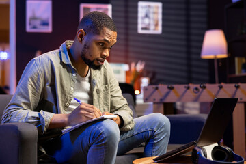 Young adult does his research by reading articles and taking notes on sofa in his living room,...