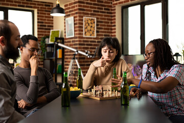 In brick wall room, group of multiethnic friends seated around table having a chess match moment. Diverse male and female individuals with drinks and snacks, concentrating on casual board game.