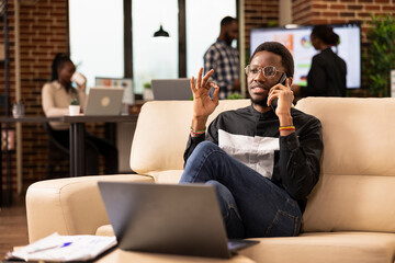 Black male professional in smart casual clothing, sitting on couch and having an important phone conversation with potential client. African american businessman using smartphone in modern workspace.