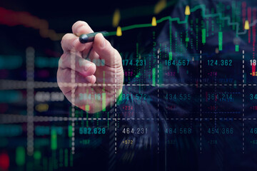 A close-up of a hand with a stylus interacting with a digital stock market chart. The overlay of candlestick graphs, buy and sell signals and numerical data.