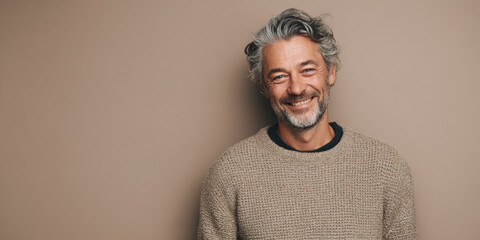 Portrait of a man with salt-and-pepper hair and a warm smile, wearing a casual sweater, standing against a neutral taupe background, relaxed and approachable