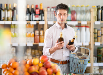 Man in a European supermarket chooses craft beer in dark glass bottles against the background of food shelves