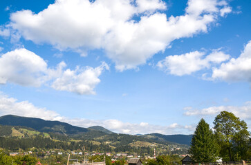 Scenic view showcasing picturesque mountain terrain, lush green trees, and scattered houses beneath a bright blue sky with fluffy cloud. Verkhovyna, Carpathian Mountains, Ukraine