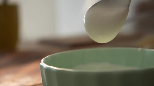 Spoon lifting and dropping thick white yogurt above a green bowl
