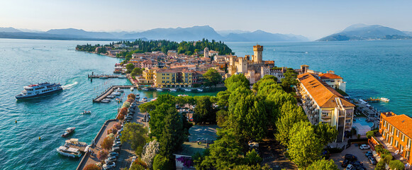Naklejka premium Aerial view of Sirmione, Italy, with boats, historic Scaliger Castle, waterfront promenade, and Lake Garda surrounded by mountains