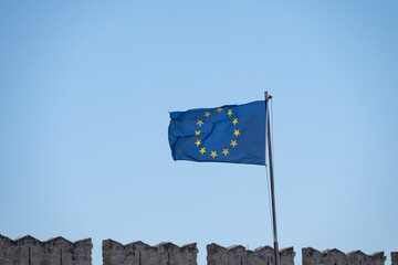 The European Union flag waving against a clear blue sky, with part of a historic stone fortress...