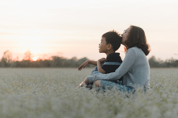 A loving mother embraces her young son as they sit together in peaceful flower field during sunset. Enjoying quiet affectionate moment in nature. Family love, motherhood, outdoor relaxation, lifestyle