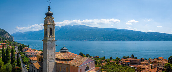 Aerial view of a pink historic church with bell tower in Gargnano, Lake Garda, Italy, surrounded by mountains and blue lake waters.