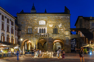 Obraz premium Night view of Bergamo’s Contarini fountain in Italy with terracotta rooftops, historic churches, and mountain backdrop on a clear day
