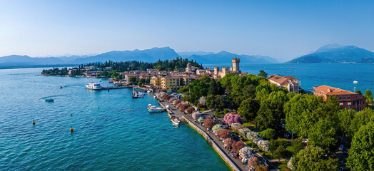 Aerial view of Sirmione, Italy, with boats, historic Scaliger Castle, waterfront promenade, and Lake Garda surrounded by mountains