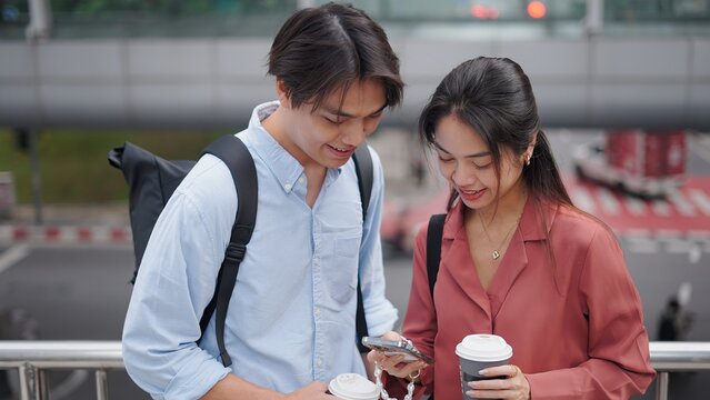 Two young professionals are using a smartphone and holding coffee while standing on a footbridge in the city - Powered by Adobe