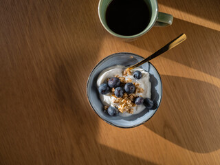 Greek yogurt with fresh blueberries and granola in blue ceramic bowl beside black coffee on wooden table