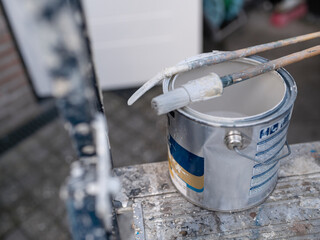 White paint bucket with brush resting on rim and ladder in background during painting project © Victor