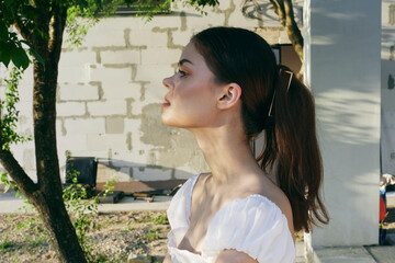 Beautiful young model with a ponytail, wearing a stylish white dress against a natural outdoor...