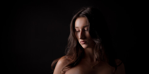 Studio portrait of a young Caucasian woman with long brown hair looking down, illuminated by a soft light against a black background
