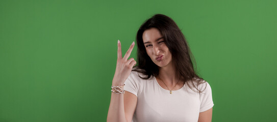 Young woman showing victory sign with fingers and making a funny face on green background